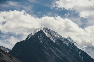 Atmosferik alp manzarası, yüksek dağ silueti, tepede mavi bulutlu gökyüzünün altında karla kaplı. Karla kaplı, sivri uçlu ve yüksek karlı tepeli dramatik dağ manzarası..