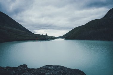 Karanlık zamanlarda bulutlu gökyüzünün altında, dağlık vadide, dağ gölü yakınlarında turuncu çadırı olan karanlık atmosferik manzara. Alacakaranlıkta dağ gölü suyunda dalgalanmalar. Highland Gölü 'nde su dalgalanması olan karanlık manzara