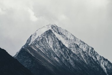 Bulutlu gökyüzünde karla kaplı yüksek siyah kayalık dağ tepeli karanlık atmosferik manzara. Karla kaplı dramatik dağ manzarası bulutlu havada sislerle kaplandı. Sisteki muhteşem kayaların kasvetli manzarası