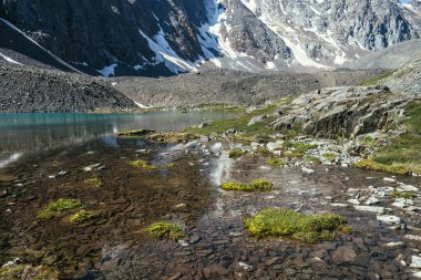 Turkuaz dağ gölü, şeffaf su ve taştan tabanlı güzel manzara manzarası. Güneş ışığında berrak su yüzeyi olan gök mavisi bir buzul gölü. Dağ gölü yakınlarında beyaz kar ve yeşil çimen.