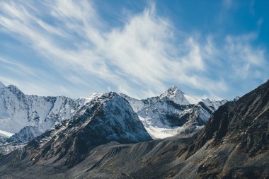 Yüksek karlı dağları olan dağlık bir arazi. Gökyüzünde sirrus bulutlarının altında zirveye ulaşmış. Güneş ışığıyla kaplı dağlara atmosferik bir manzara. Siyah kayalar ve beyaz kar, gün ışığında sivri tepeler..
