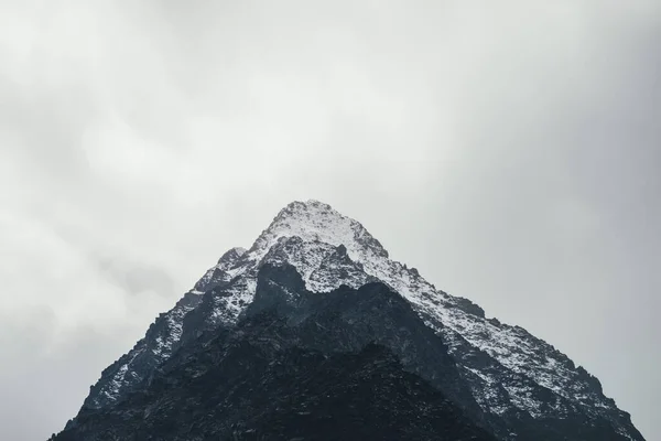 曇り空の雪と高い黒い岩の山の頂上と暗い大気の風景 雪の劇的な山の風景は曇りの天気で霞の上にピーク 霧の中で素晴らしい岩への悲観的な眺め — ストック写真