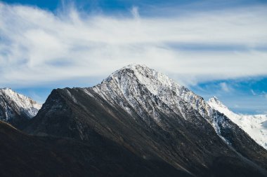 Atmosferik alp manzarası. Mavi gökyüzünde, sirrus bulutlarının altında, karla kaplı kayalık dağın tepesi. Harika bir dağ manzarası. Sivri uçlu, karlı, güzel bir silueti var. Büyük kayalık zirve.