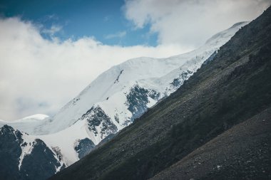 Karlı dağ sıraları ve bulutlu gökyüzünün altında çapraz dik yamaçları olan muhteşem dağlar. Atmosferik dağ manzarası. Kar beyazı yüksek dağ sırtı ve bulutların altındaki kayalık dağ yamacı..