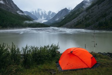 Atmosferik alp manzarası, yeşil dağ gölü kıyısında turuncu çadır ve yağmurlu havada karlı dağlar. Dağ gölünün suları üzerinde yağmurlu daireler ve vadideki alçak bulutlarla kasvetli bir manzara.