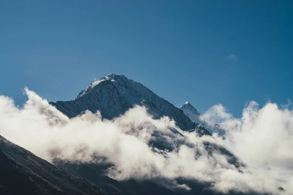 Minimalist view of snow-capped mountain silhouette above thick clouds. Scenic mountain landscape with white-snow sharp peak among dense low clouds in blue sky. Wonderful scenery with snowy pinnacle.