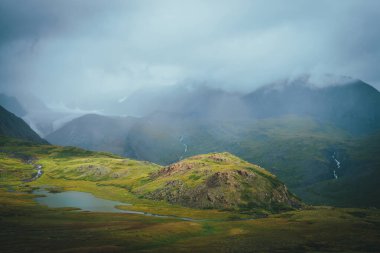 Yeşil vadideki güzel dağ gölü, büyük kayalar ve bulutlu havada yüksek dağların arasında dramatik alp manzarası. Göl manzarası ve alçak bulutlarda buzul manzarası. Atmosferik manzara.