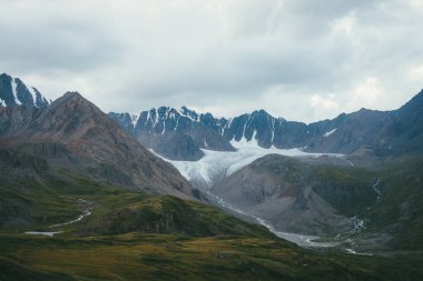 Bulutlu bir havada vadide büyük bir buzul ve dağ nehri ile dramatik bir alp manzarası. Bulutlu gökyüzünün altındaki dağlık atmosfer manzarası. Gri gökyüzünün altındaki güzel dağ sırtı.