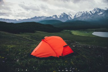 Atmosferik alp manzarası gölün arka planında turuncu çadır ve bulutlu havada büyük karlı dağlar. Turuncu çadırlı, bulutlu gökyüzünün altındaki büyük dağların manzaralı muhteşem yeşil manzarası..