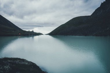 Karanlık zamanlarda bulutlu gökyüzünün altında, dağlık vadide, dağ gölü yakınlarında turuncu çadırı olan karanlık atmosferik manzara. Alacakaranlıkta dağ gölü suyunda dalgalanmalar. Highland Gölü 'nde su dalgalanması olan karanlık manzara