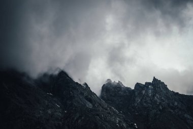 Gri bulutlu gökyüzünde siyah kayalık dağ duvarlı karanlık atmosferik manzara. Kara dağın tepesinde gri alçak bulut. Karanlık dağ zirvesi alçak bulutlarda bulutlu havada. Kasvetli minimalist manzara
