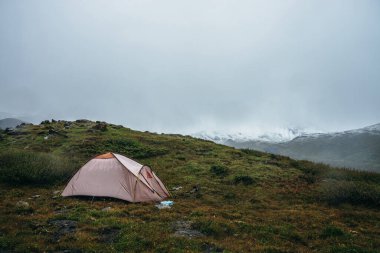 Yeşil tepelerde kayaların arasında çadırları olan manzaralı bir dağ manzarası. Alçak bulutlarda kar kaplı güzel dağlar. Dağlık bölgelerde, bulutlu havalarda çadırlı atmosferik manzara. Yüksek dağlar karla kaplı.