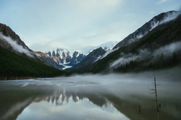 低雲の間で霧の中でミラー山の湖に反映黄金の太陽の下で雪の山と大気高山の風景 岩や緑の鏡湖の上に低い雲と風景高原の風景 — ストック写真