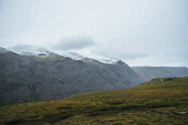 Karlı dağlarla kaplı minimalist dağlarda alçak bulutlar ve bulutlu hava. Düşük bulutlu gökyüzünün altındaki karlı dağ sırasının arka planında derin bir vadi. Yüksek dağlarda atmosfer minimalizmi.