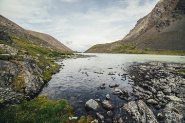 Bulutlu gökyüzünün altında, ufuktaki dağların arkasındaki dağ gölüne güzel bir manzara. Yüksek dağ vadisinde gölü olan atmosferik yeşil bir manzara. Dağlık vadide dağ gölü manzarası