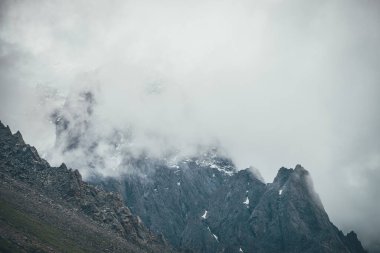 Karanlık atmosferik gerçeküstü manzara. Koyu kayalık dağ tepesi, alçak bulutlar ve gri bulutlu gökyüzü. Yüksek zirvede gri alçak bulut. Alçak bulutlarda kar olan yüksek siyah kaya. Sürrealist kasvetli dağlar.