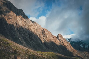 Alçak bulutlardaki yüksek karlı dağların arka planında güneşli kayalık bir tepe noktası olan muhteşem bir manzara. Keskin kayaları ve bulutlu gökyüzünde dev karlı dağları olan Atmosferik Alp manzarası. Manzaralı alp görünümü.