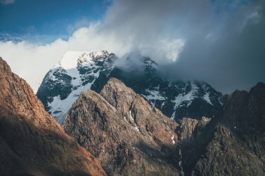 Büyük kayaların ve karlı dağların olduğu manzaralı alçak bulutlarda güneş ışığı alan bir yer. Bulutlu gökyüzünde karla kaplı dağın zirvesine harika bir manzara. Güneşli, kar beyazı tepeli muhteşem bir manzara..