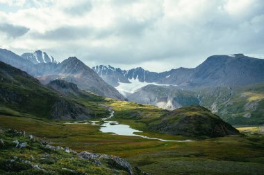 Atmosferik alp manzarası, yeşil vadide dağ gölü ve bulutlu gökyüzünün altında buzul. Güneşli tepeler ve dağ sırasına karşı kayalar arasında buzul gölü olan muhteşem dağlık arazi manzarası..