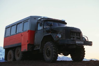 Tepedeki turuncu kamyona yakın. Gün batımında gökyüzünün altındaki büyük kar dağlarına bakıyor. Kamyon, parlak gün ışığında karlı dağların arka planına yaklaşıyor. Rusya, Altai Cumhuriyeti, 20 Eylül 2020.