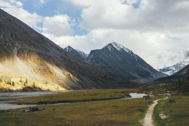Sonbahar renklerinde vadideki su akarsuları boyunca uzanan, karlı dağları ve altın güneş ışığındaki kayaları gören manzaralı bir manzara. Dağ deresi ve karlı dağları olan renkli sonbahar manzarası