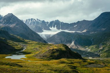 Atmosferik alp manzarası, yeşil vadide dağ gölü ve bulutlu gökyüzünün altında buzul. Güneşli tepeler ve dağ sırasına karşı kayalar arasında buzul gölü olan muhteşem dağlık arazi manzarası..