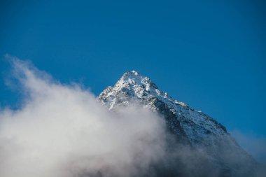 Kalın bulutların üzerinde, gün ışığında, keskin karla kaplı dağ manzarası harika. Mavi gökyüzündeki yoğun alçak bulutların arasında beyaz kar zirvesi olan parlak dağ manzarası. Karlı zirvesi olan en küçük manzara.
