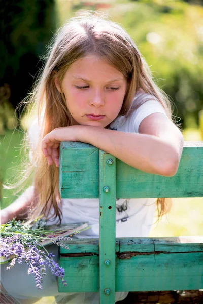 Η σκεπτική κοπέλα κάθεται σε ένα παγκάκι με ένα μπουκέτο λουλούδια κοντά σε ένα ιδιωτικό σπίτι. — Φωτογραφία Αρχείου
