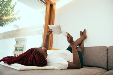 Siyahi kadın kanepede uzanıp ayaklarını duvara dayayıp kitap okuyor.