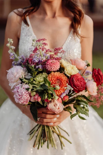 Wedding bouquet of red, orange and purple flowers close-up in the hands of a bride in a white wedding dress at sunset with warm color
