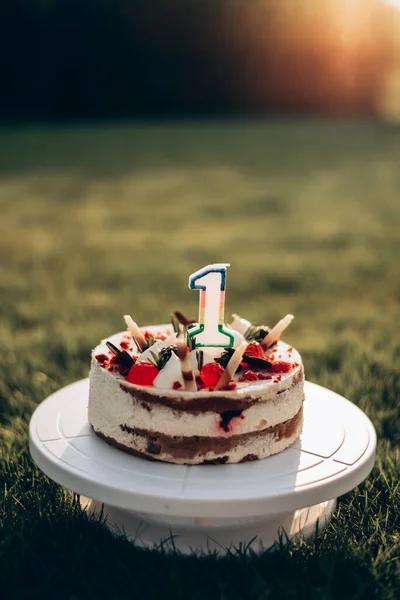 Cake with fresh raspberries and blackberries for a child\'s birthday with a candle in the form of the number one. Cake stands on a green lawn, at sunset with a blurred background
