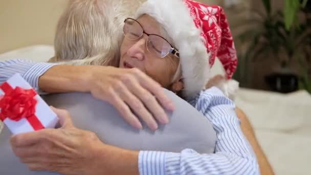 Счастливая пожилая пара обменивается рождественскими подарками дома. пожилой мужчина дарит пожилой женщине коробку с рождественским декором, а женщина с благодарностью обнимает мужчину. Рождественское утро — стоковое видео