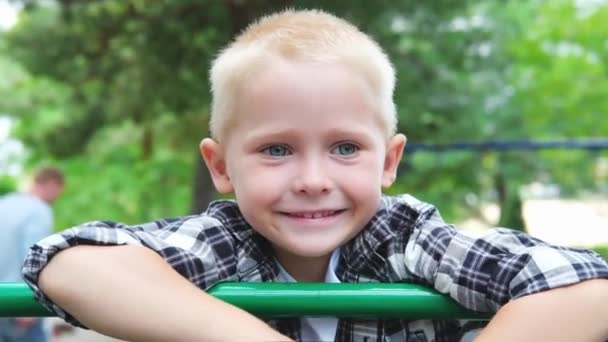 Portret drăguț băiat blond păr cu ochi albaștri care se joacă pe terenul de joacă. Copii zâmbind și uitându-se la camera din parc la apusul soarelui. Conceptul de copil vis de familie fericit. mișcare lentă — Videoclip de stoc