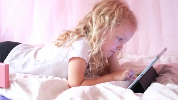 Кавказька допитлива кучерява дівчинка дошкільного віку використовує цифровий пристрій для планшетів, що лежить на ліжку сам. Маленька дитина проводить домашню інтернет-гру серфінгу. Діти - технологи — стокове відео