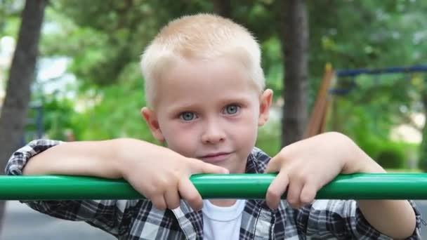 Portrét roztomilé blond vlasy chlapec s modrýma očima hraje na hřišti. děti se usmívají a dívají se na kameru v parku při západu slunce. Šťastný rodinný sen dítě koncept. zpomalený pohyb — Stock video