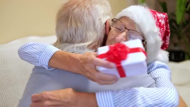 Fermer portrait Heureux couple de personnes âgées échangeant des cadeaux de Noël à la maison. homme âgé donne à une femme âgée une boîte avec décor de Noël et Femme étreint avec gratitude un homme. Matin de Noël en famille — Video