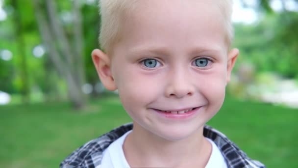 Retrato ao ar livre bonito de um menino branco branco de olhos azuis sorridentes. O menino gosta de close-up. criança no parque de verão. criança sorri amplamente com dentes brancos — Vídeo de Stock