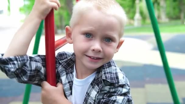 遊び場で遊んでいる青い目のかわいいブロンドの髪の男の子の肖像画。子供たちは笑顔で公園でカメラを見ています。幸せな家族の夢の子供の概念。スローモーション — ストック動画