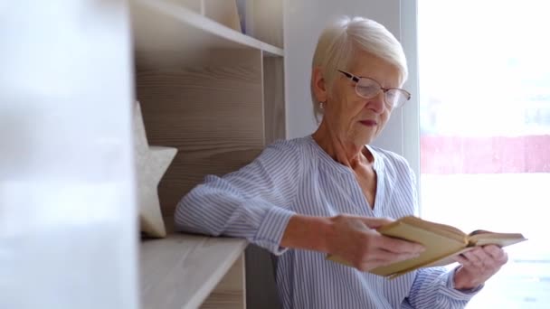 在家里的图书馆里，戴眼镜的高年级的高加索女人正在看一本古老的历史书。成年女性在舒适的现代公寓中的休闲活动.临时的老年生活。4.退休的退休人员喜欢看书 — 图库视频影像