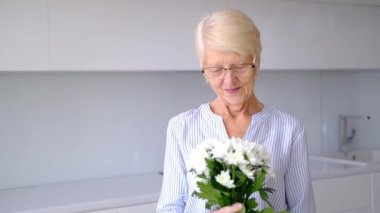 Güzel, yaşlı, mutlu, yaşlı, beyaz bir kadın çiçek buketi koklarken kameraya bakıp gülümsüyor. Mutfak odasındaki mutlu yaşlı kadın portresi. Kadınlar Günü 'nde elinde çiçeklerle poz veren kadın..