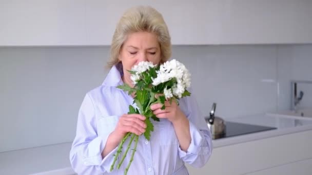 Krásná šťastná dospělá běloška šťastná vonící kytice květin při pohledu na fotoaparát s úsměvem. portrét šťastné ženy v kuchyňském pokoji uvnitř. Dáma pózuje s květinami na den žen. — Stock video