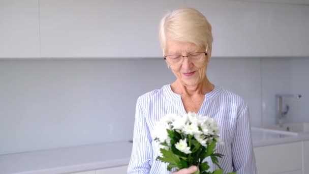Beautiful old happy senior Caucasian woman smelling bouquet of flowers looking at camera smiling. portrait of happy elderly woman in kitchen room indoors. lady posing with flowers on Womens Day. — Video Stock