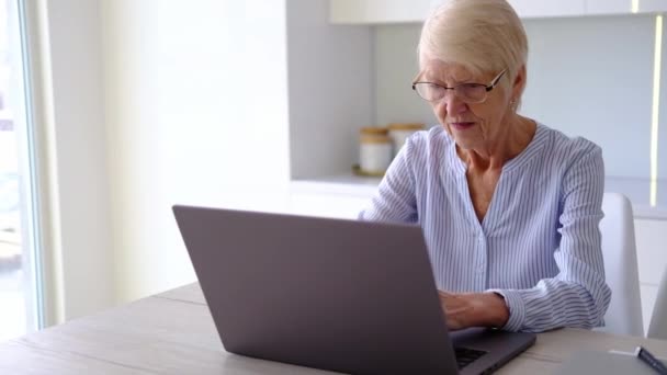 Senior mujer mayor madura que trabaja en el ordenador portátil en la casa de la cocina en la oficina. señora viendo formación de negocios, webinar en línea en el ordenador. aprendizaje a distancia social de trabajo remoto desde el hogar. Mujer de 60 años — Vídeo de stock