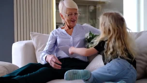 Happy cute curly little child granddaughter jump on sofa and embracing kissing old aged grandmother. girl presenting bouquet flower for old lady making surprise for birthday , hugging having fun — Stock Video