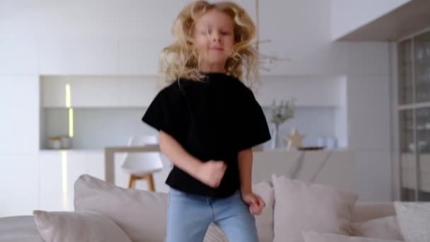 Retrato bonito bonito menina loira encaracolado vestindo uma camiseta preta e jeans rindo pulando no sofá com almofadas na sala de estar. sozinho feliz adorável criança se divertindo na cama. — Vídeo de Stock