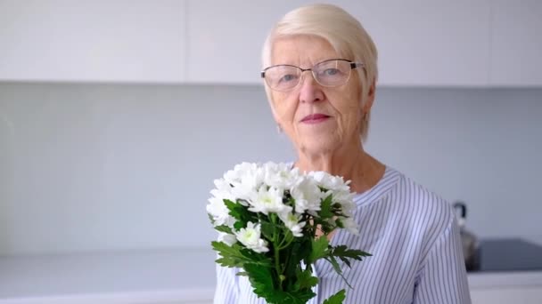 Vacker gammal glad senior vit kvinna luktar bukett blommor tittar på kameran ler. Porträtt av lycklig äldre kvinna i köksrummet inomhus. dam poserar med blommor på kvinnodagen. — Stockvideo