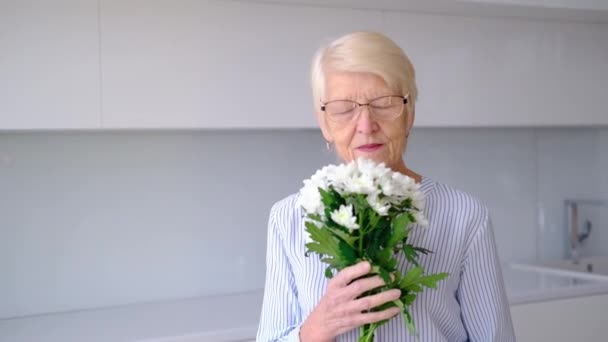 Beautiful old happy senior Caucasian woman smelling bouquet of flowers looking at camera smiling. portrait of happy elderly woman in kitchen room indoors. lady posing with flowers on Womens Day. — Stock Video