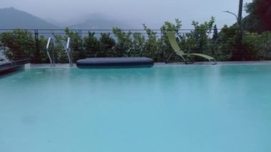 Yağmur damlaları Como Gölü, Lombardy, İtalya 'da bir yüzme havuzuna düşüyor. Bulutlu, bulutlu, yağmurlu bir yaz günü. Havuzun arkasında bir şişme yatak ve bir güneş sandalyesi var. Arka planda gölü çevreleyen dağlar var..