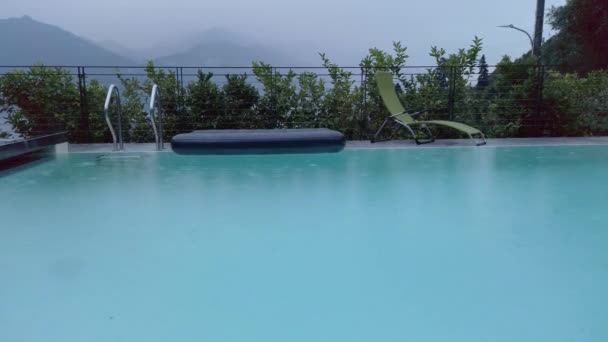 雨滴落在意大利伦巴第科莫湖畔的一个游泳池里 这是一个阴云密布 阴沉沉 多雨的夏日 游泳池后面有一个气垫和一把太阳能椅 背景上有群山环绕着湖面 — 图库视频影像