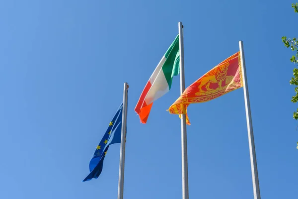 Italian, EU and the local flag of the Italian Veneto region waving in the wind. It is a beautiful sunny summer day, with a clear blue sky in the background.